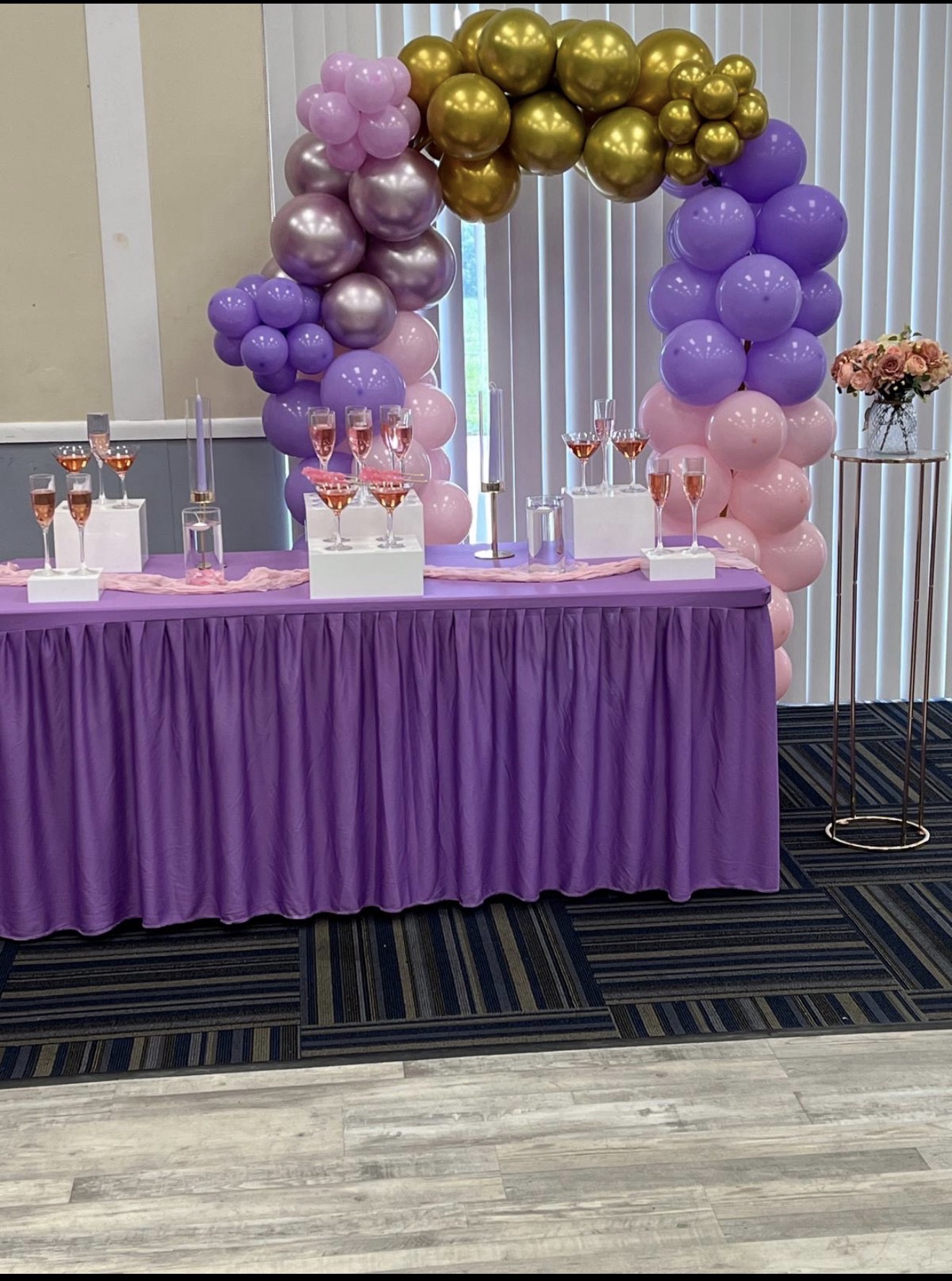 Purple, pink and gold balloon arch over a welcome table of champagne glasses